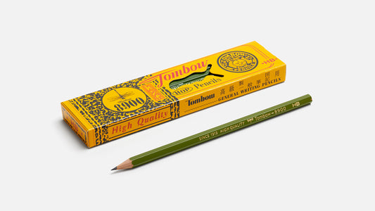 A green wooden pencil with gold lettering lies diagonally in front of its yellow and black packaging box, which features the brand name 'Tombow' and 'General Bating Pencil'.
