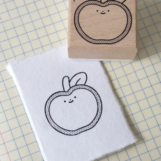 A small white fabric square with a black outlined cute apple face, shown next to a wooden rubber stamp block with the same apple design on a beige graph-paper background.