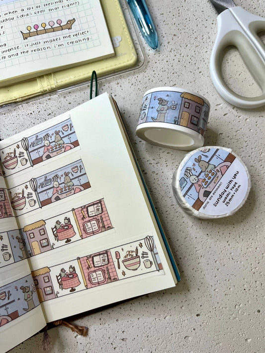 Open notebook with illustrated pages, a small container, and a roll of decorative tape on a textured surface.