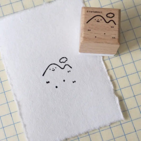 A wooden rubber stamp with a simple mountain and sun illustration sits on a sheet of white paper that has been stamped with the same design. The stamp is placed on a pale yellow grid mat.