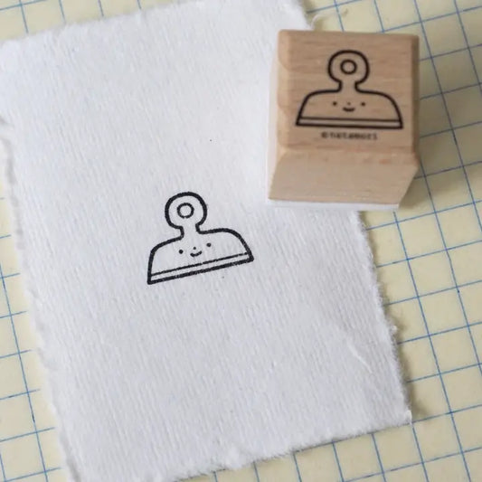 A wooden rubber stamp with a simple smiling clipboard or clipboard-like character design, shown next to a white textured sheet that has been stamped with the same black outline.