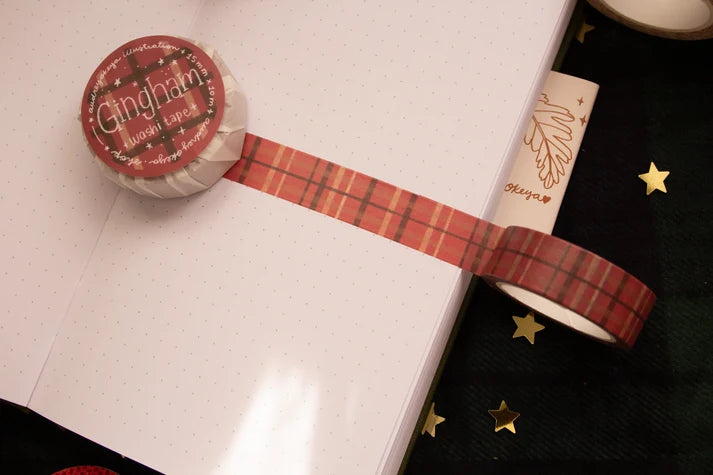 A roll of Gingham washi tape with a red and orange plaid pattern, laid across a white notebook page next to another roll of washi tape on a black starry background.
