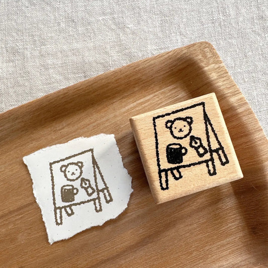 Wooden stamp with a design of a bear holding a mug and a piece of paper with the same design on a wooden tray.