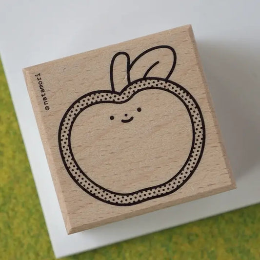 A square wooden rubber stamp with a cute, minimalist apple design featuring a smiling face and dotted border. The stamp is shown resting on a white card over a textured green and yellow surface.