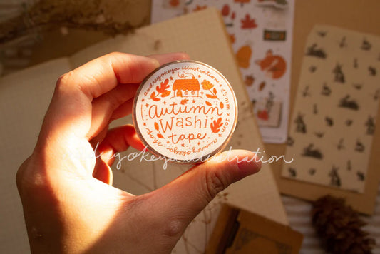 Hand holding a container of autumn-themed washi tape with decorative elements.