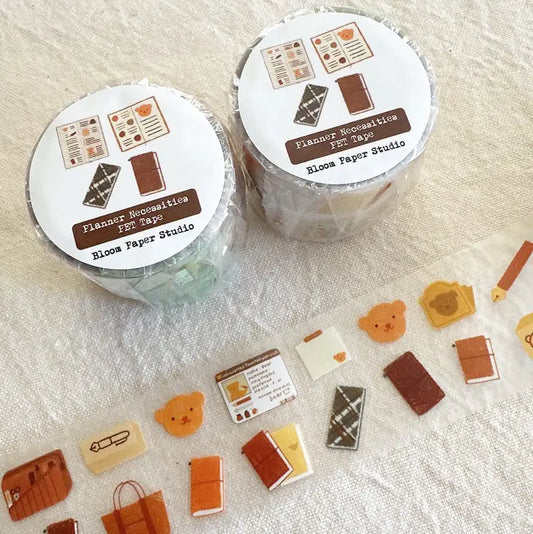 Two rolls of Planner Necesities PET Tapes from Bloom Paper Studio on a textured surface with scattered tape shapes.