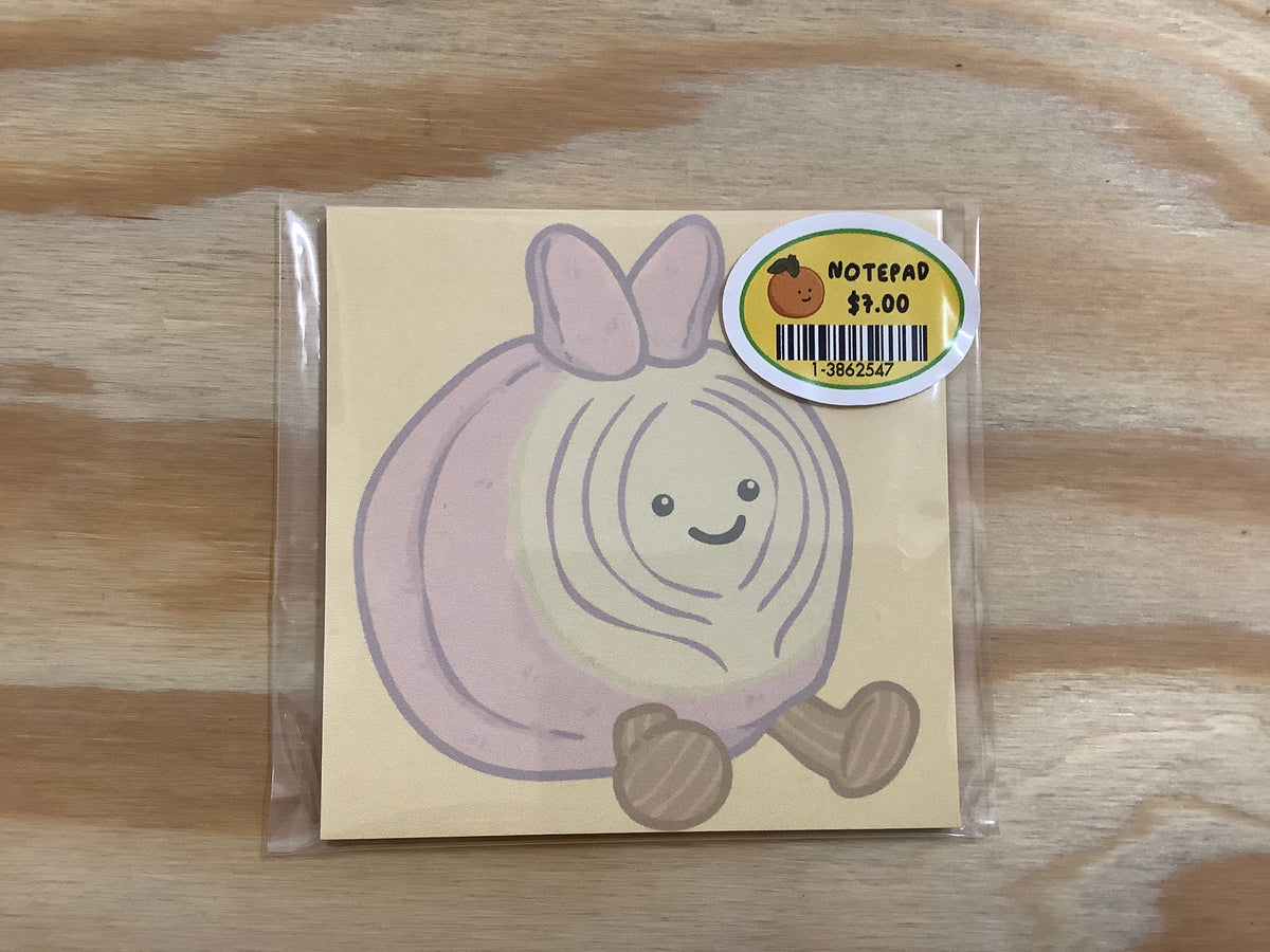 A cute onion character notepad inside a clear plastic sleeve, resting on a light wood grain surface. The notepad features a smiling cartoon onion with pinkish-purple layers, a yellow sticker showing the price of $3.
