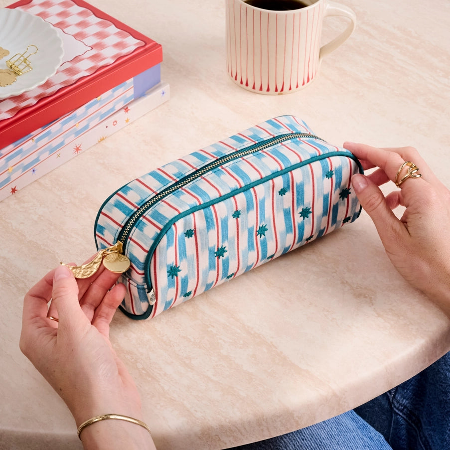 Person opening a striped pencil case on a table with a mug and stationery items in the background.