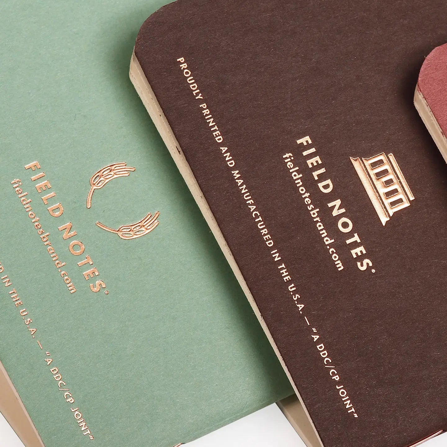 Two Field Notes notebooks, one green and one brown, with gold embossed text on a white background.