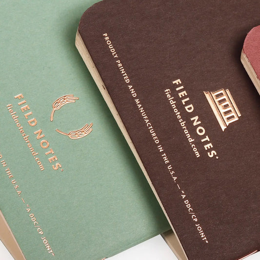 Two Field Notes notebooks, one green and one brown, with gold embossed text on a white background.