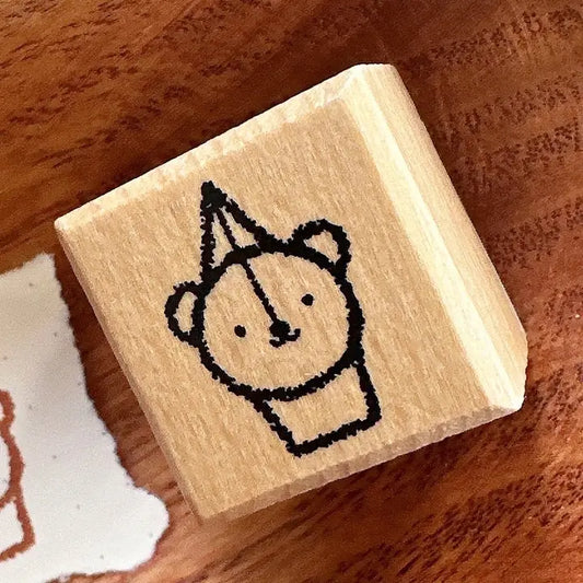 A wooden rubber stamp block featuring a simple black line drawing of a bear wearing a party hat, resting on a warm brown wooden surface.
