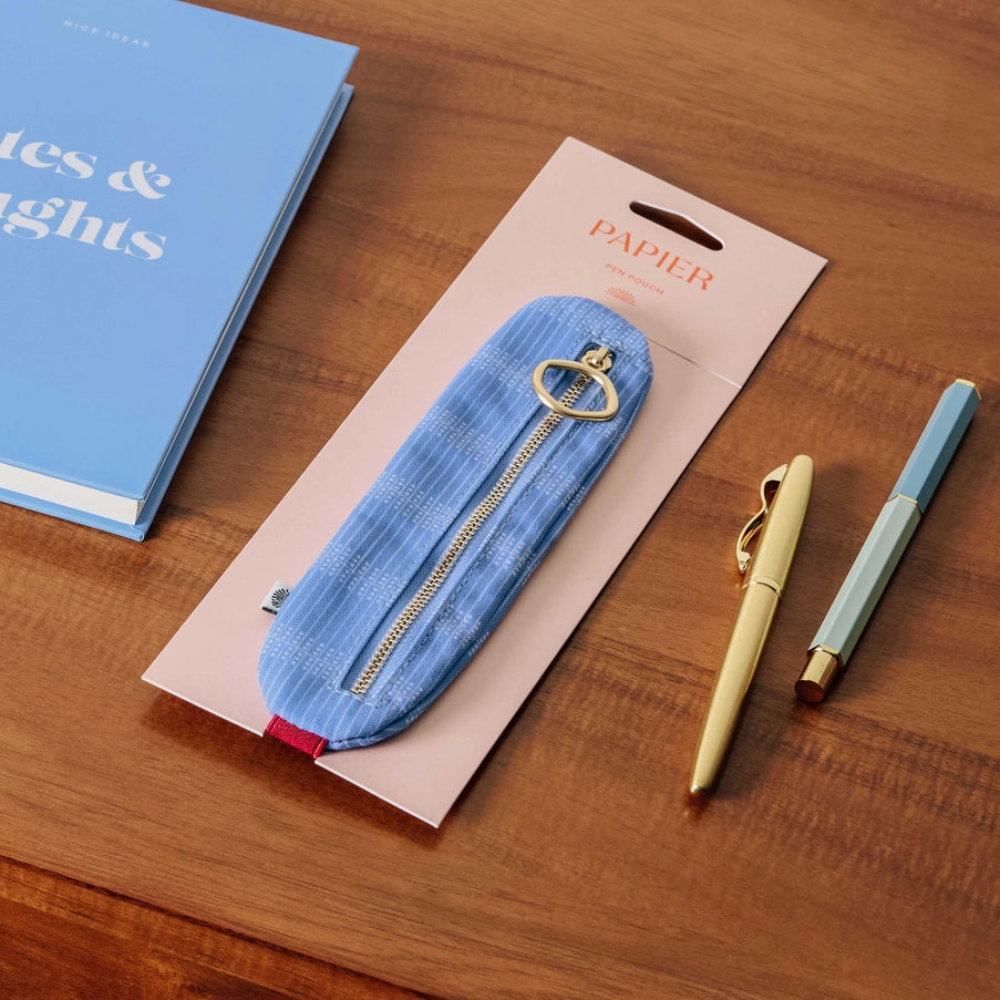 Blue denim-style pencil case with gold zipper on a wooden surface, accompanied by two pens and a notebook.