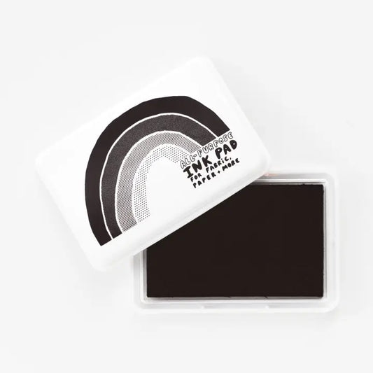 A square TNK pad ink pad with its white lid resting at an angle above the open clear plastic case, showing the dark brown ink pad inside. The lid features a black and gray rainbow graphic with the text 'After Every Meal TNK PAD THE TRADITIONAL PAPER INK'.