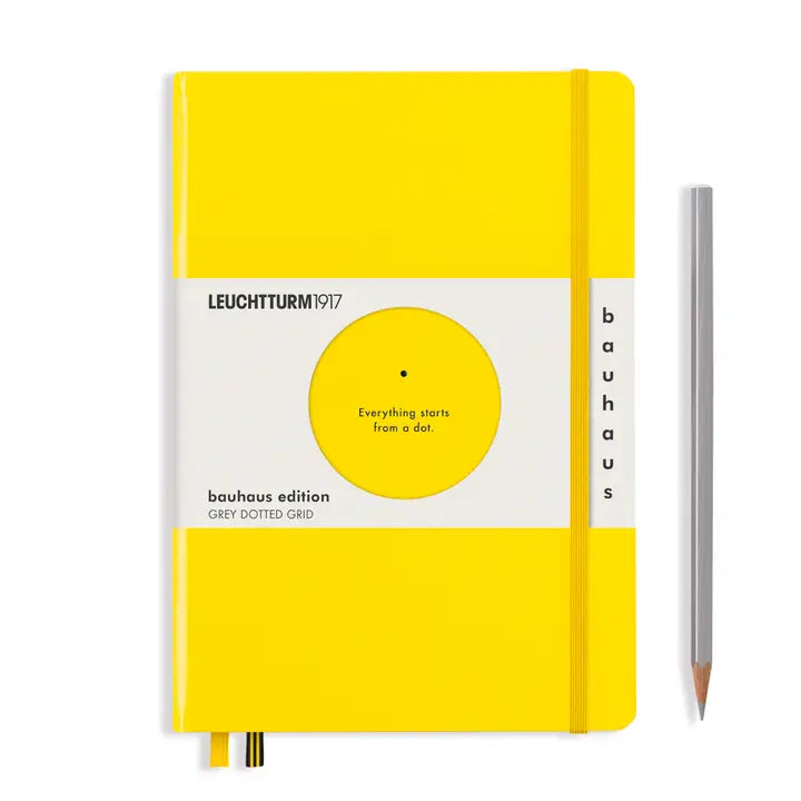 A Leuchtturm1917 bauhaus edition grey dotted grid notebook in bright yellow with a white cover band, shown upright next to a silver pencil on a white background.