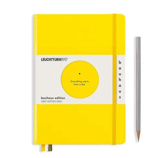 A Leuchtturm1917 bauhaus edition grey dotted grid notebook in bright yellow with a white cover band, shown upright next to a silver pencil on a white background.