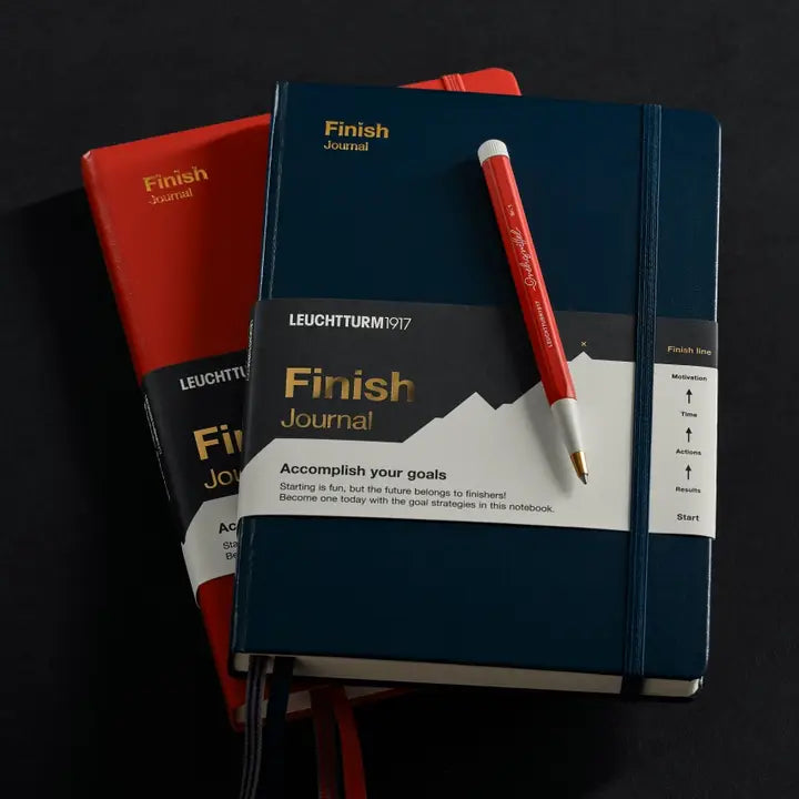 A stack of Leuchtturm1917 Finish Journal notebooks in navy blue and red, with a red and white pen resting on the top navy journal against a dark background.