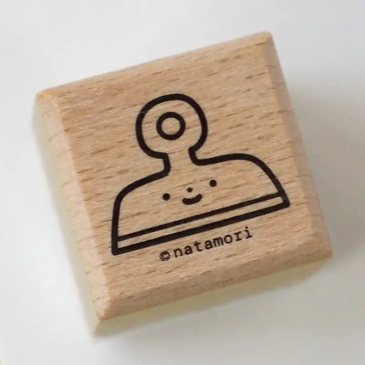A square wooden rubber stamp with a black line drawing of a smiling clipboard character and the text '@natamori' on a plain white background.
