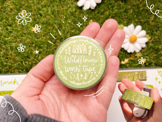 Hand holding a container of Wildflower Washi Tape with decorative elements on a grassy background