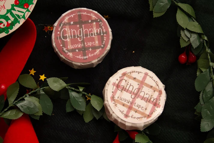 Two round packages of Gingham washi tape displayed on a dark textured background with festive Christmas decorations including green leaves, red berries, and a red ribbon.