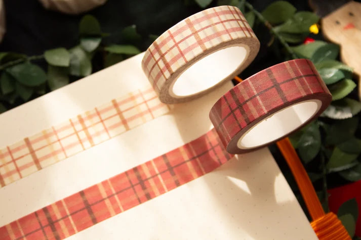 Two rolls of plaid washi tape, one in beige and orange and one in red and orange, displayed on a sheet of white paper with green foliage in the background.