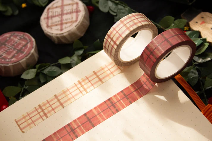 Two rolls of plaid washi tape, one in beige and red and one in red and brown, displayed on a notebook with green leaves in the background.