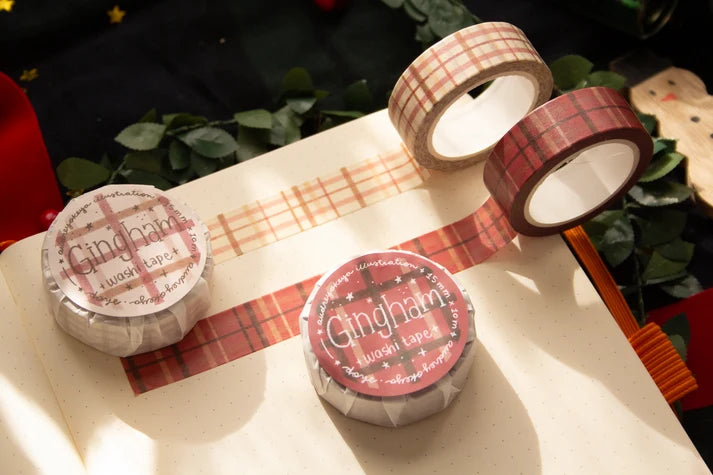 Three rolls of Gingham washi tape in red, beige, and brown plaid patterns, displayed on an open notebook with warm sunlight and green leaves in the background.