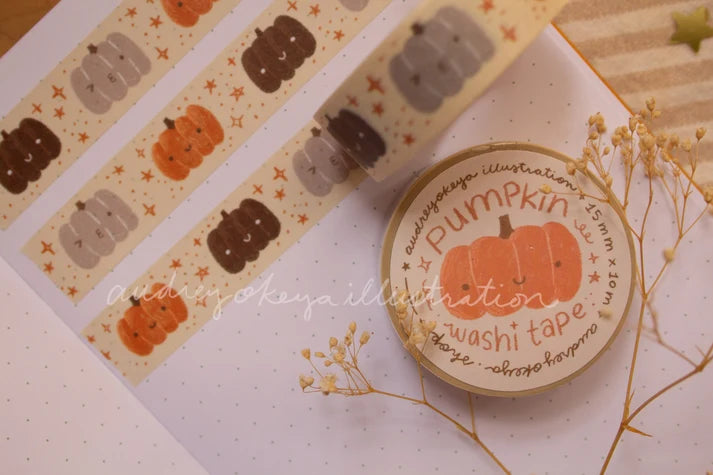 Pumpkin-themed washi tape rolls displayed on a white dotted notebook with dried flowers. The tape features cute cartoon pumpkins in orange, brown, and gray with small gold stars on a cream background.