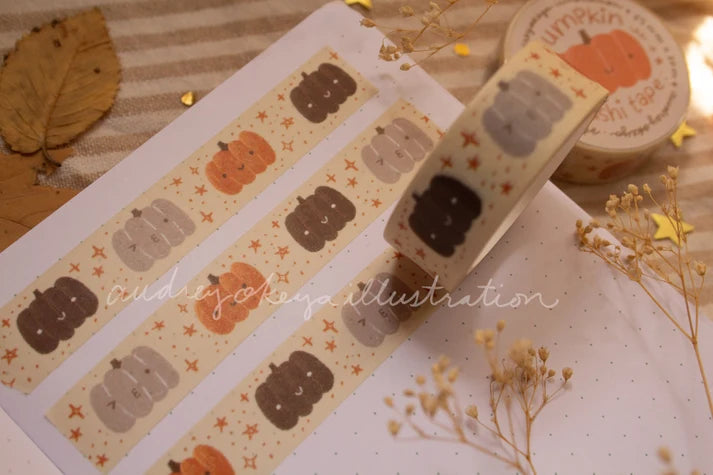 A roll of washi tape and sheets of matching washi tape featuring cute pumpkin and star illustrations, styled with dried flowers and autumn leaves on a striped fabric background.
