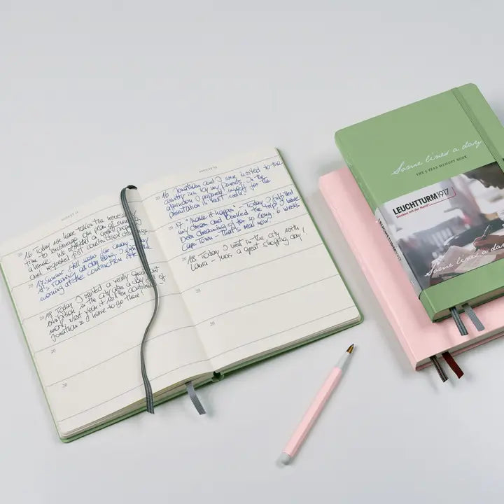 An open lined journal with handwritten notes and an elastic closure strap, shown next to a stack of notebooks in pastel pink and sage green, with a pink pen on a light gray background.