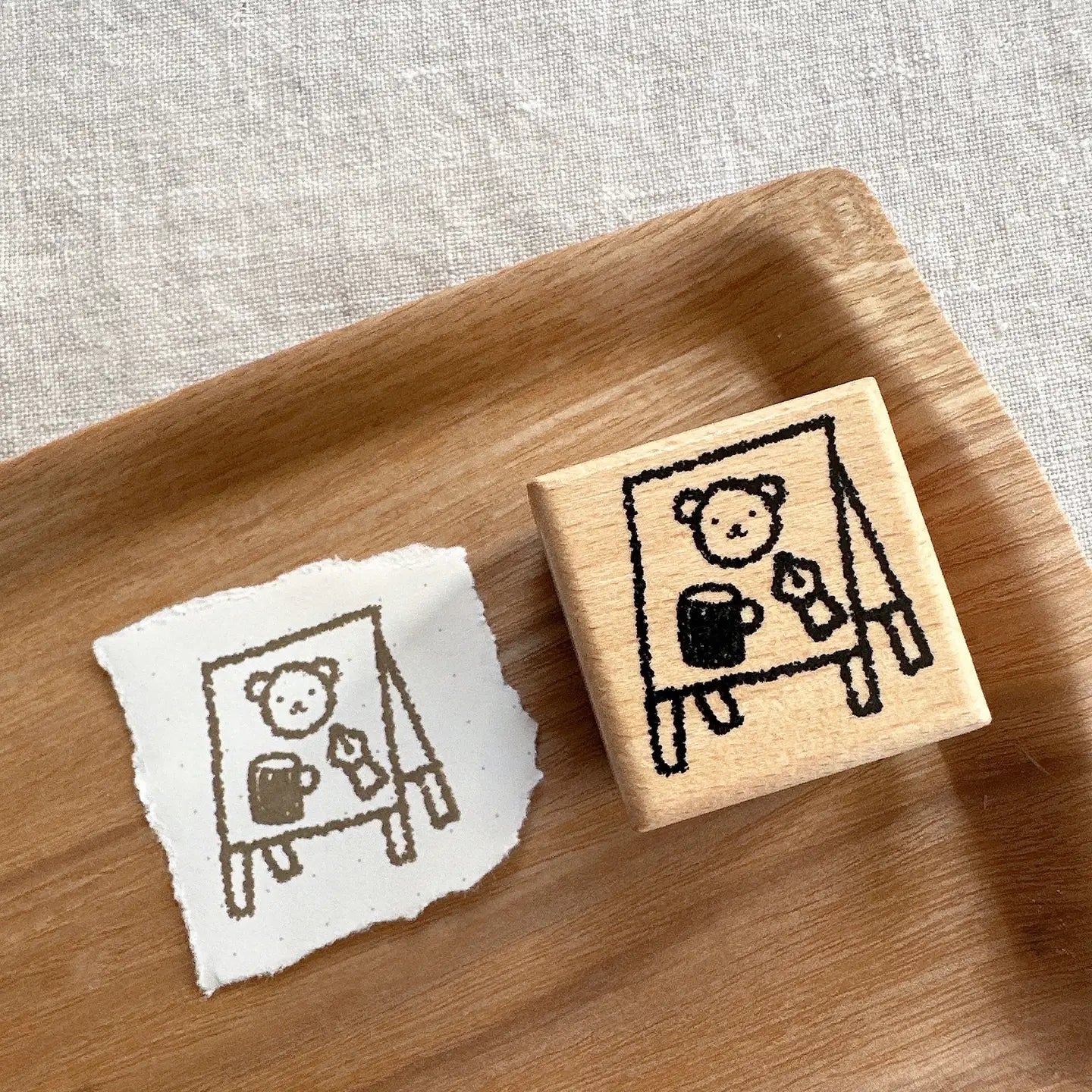 Wooden stamp with a design of a bear holding a mug and a piece of paper with the same design on a wooden tray.
