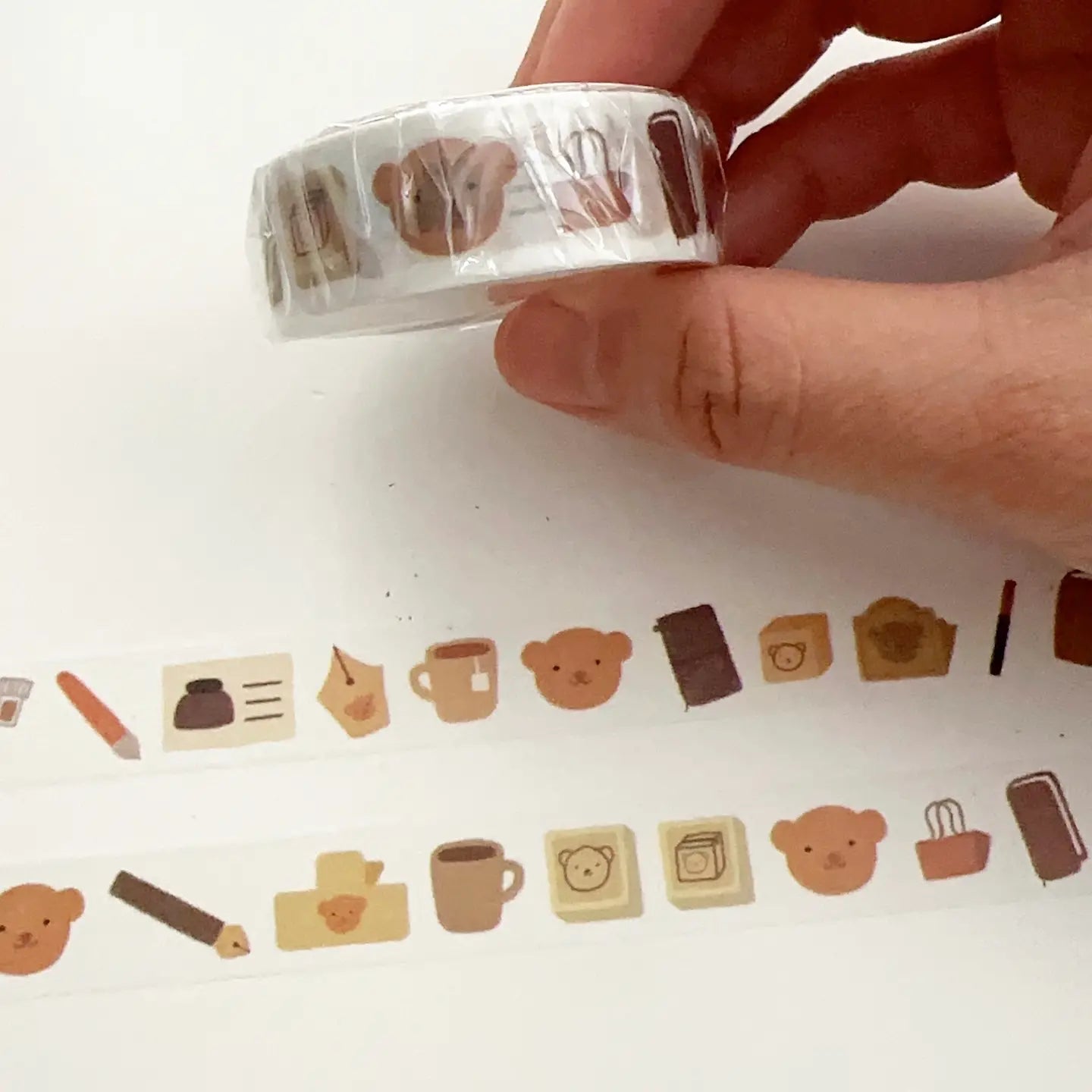 Hand holding a roll of decorative tape with small illustrations, including bears and coffee cups, on a white background.