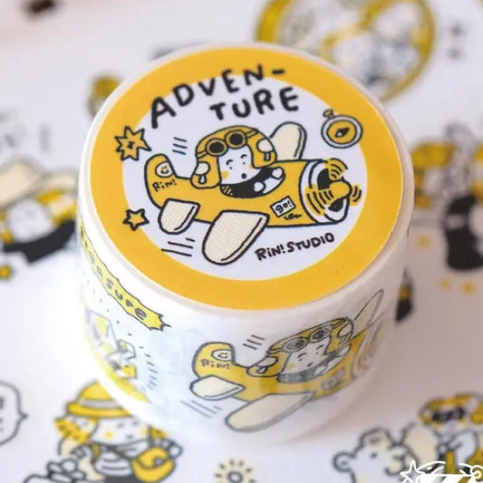A roll of washi tape featuring a yellow and white cartoon astronaut design with the word 'ADVENTURE' on the lid, placed on a matching patterned surface. The brand 'Rin: STUDIO' is visible on the lid.