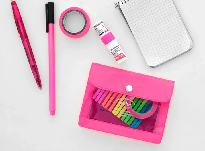 A flat-lay arrangement of pink stationery items on a white background, including a pink pen, highlighter, washi tape, small bottle, notebook, and a translucent pink pouch containing colorful sticky notes.