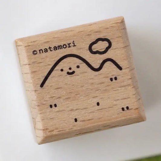 A square wooden rubber stamp block with a light natural wood grain and a black engraved design of a smiling mountain and cloud, with the text '@natamori' printed on the top left, shown on a plain white background.