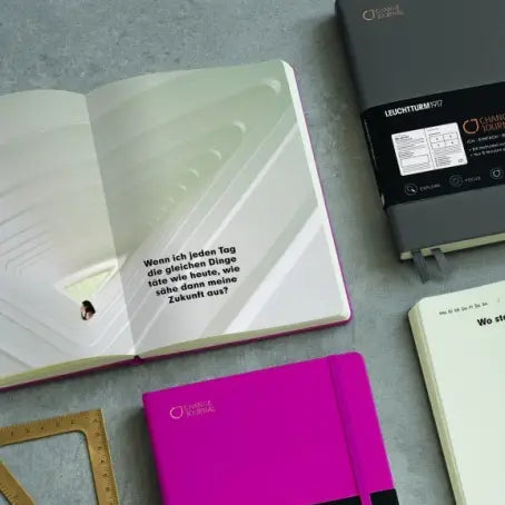 An open notebook with a white cover and German text on the left, alongside other notebooks and stationery on a gray surface. The main featured product appears to be the open notebook, with other notebooks partially visible around it.