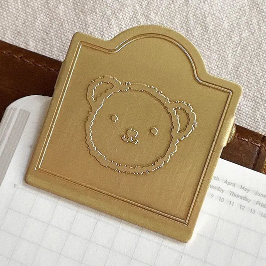 A square gold-toned metal clip or bookmark with an embossed teddy bear face, resting on a white grid notebook page.