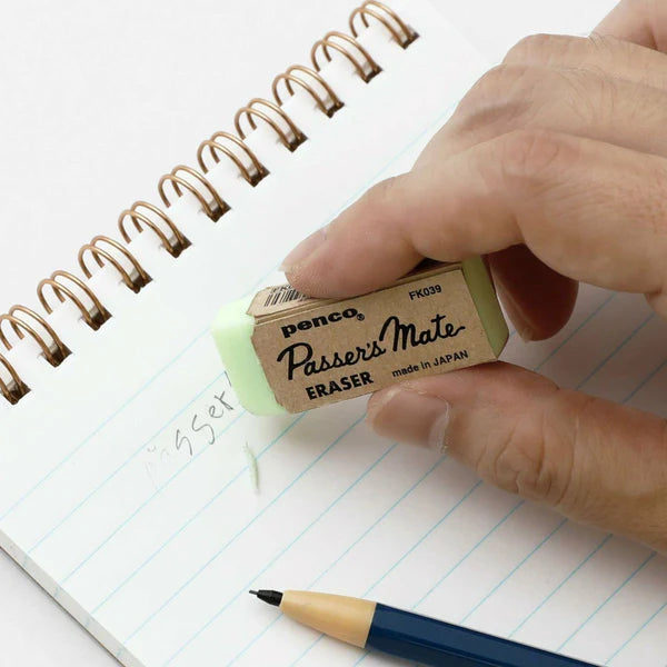 Hand holding a Passer's Mate eraser over a notebook with a pen on a white background