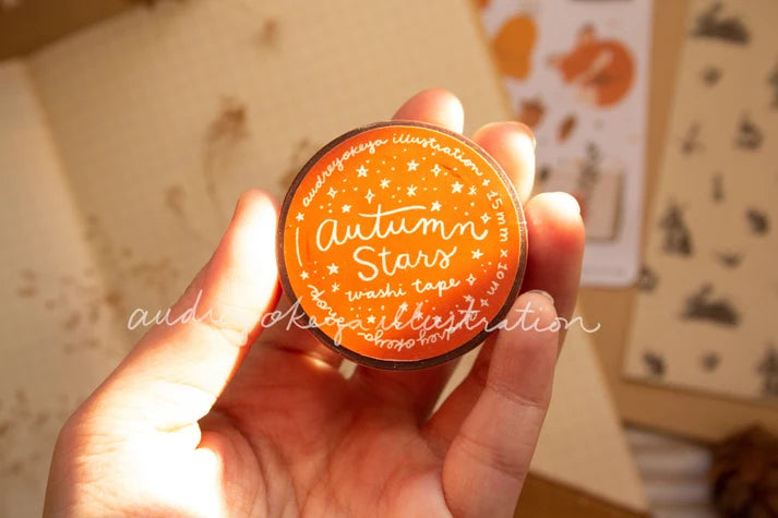 A hand holding a round orange washi tape roll labeled 'Autumn Stars' with white script and star details, set against a warm, softly blurred background.