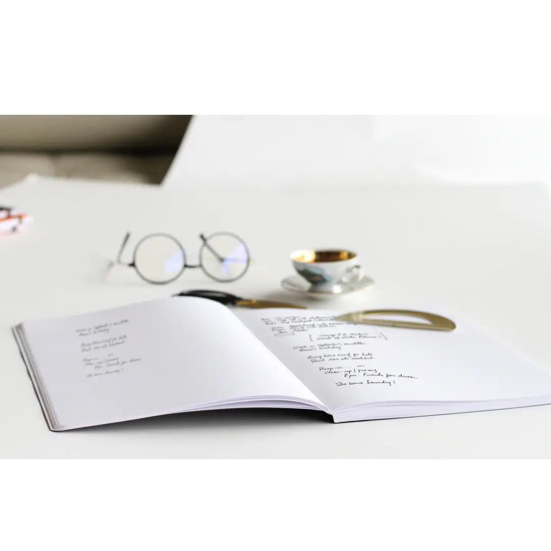 Open book on a white surface with glasses and a small cup in the background